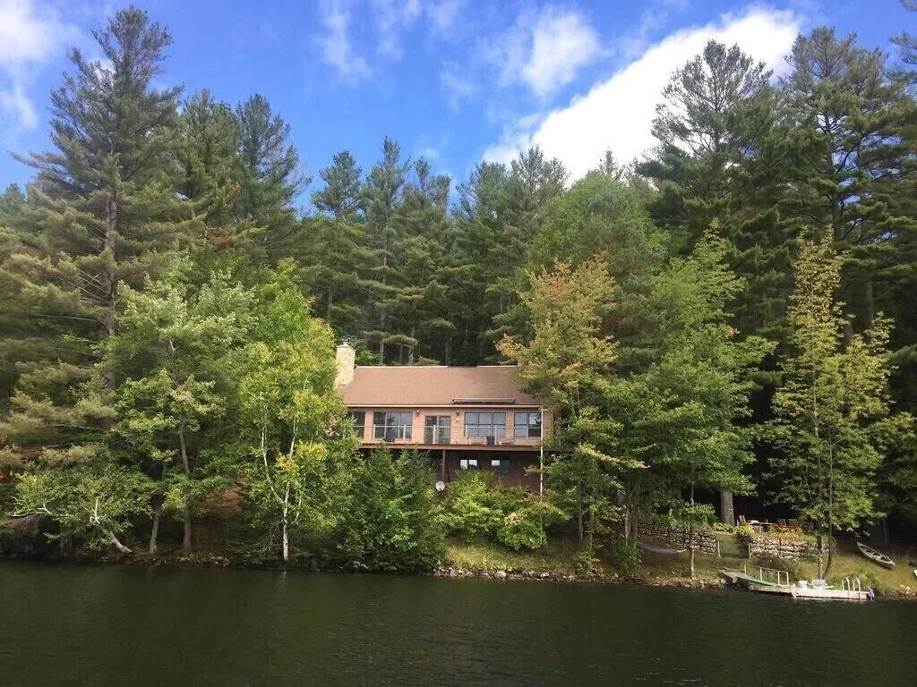 A lakeside home is nestled into a dense forest of green on the lake's edge, with a bright blue sky and white clouds visible above on a sunny day