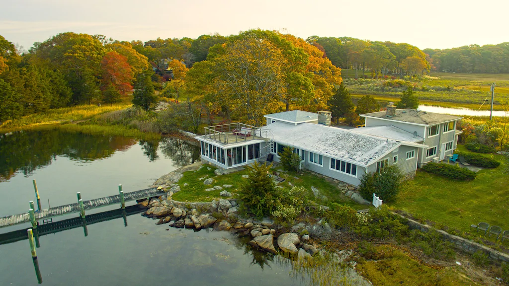 Body of water and rocky shore with a large white compound among the fall trees at a vacation rental in New England