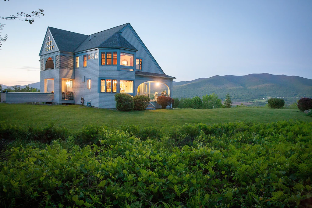A large green field in front of a beautiful New England vacation rental with interior lights lit