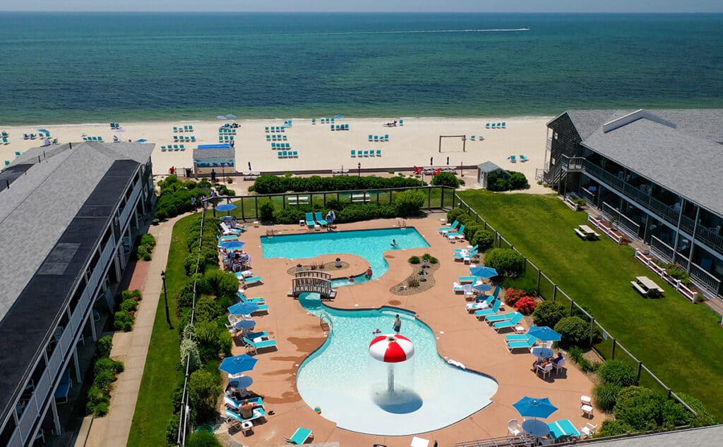 Two outdoor pools with the beach beyond are seen from an aerial point of view on Cape Cod