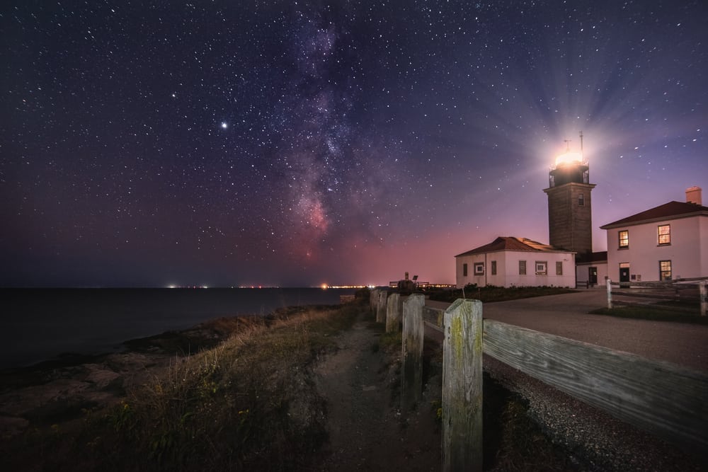 a starry night sky behind an illuminated lighthouse on the coast of Rhode Island
