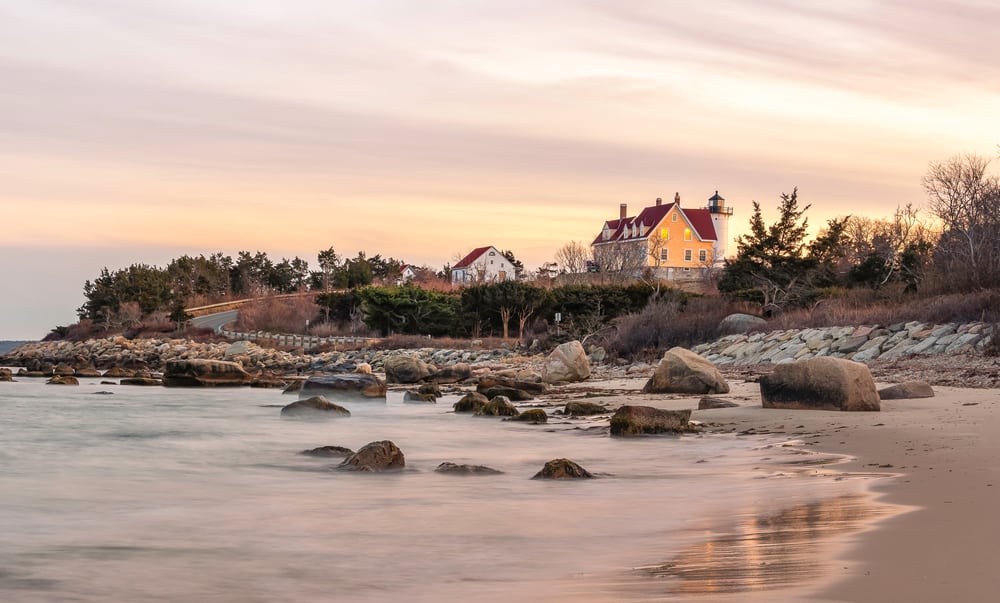 A sunset over a body of water at a rocky shore with houses in the distance