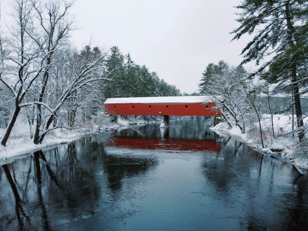 A red covered bridge with snow on top and snow on each shore with the river in between