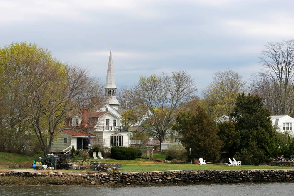 A white church with a steeple surrounded by trees with a lawn out front and a low stone wall 