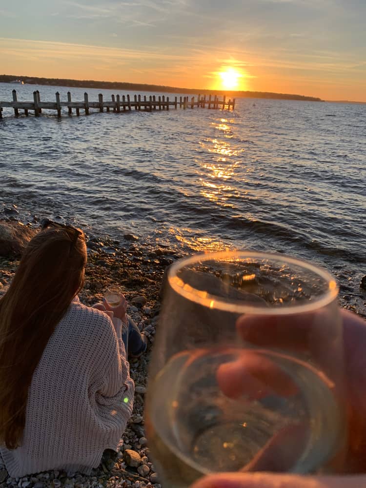 A sunset over a body of water with a closeup of a glass of wine and a girl looking out at the ocean and a pier