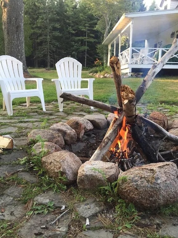 A fire pit with a fire burning next to white Adirondack chairs and a house with porch in the background