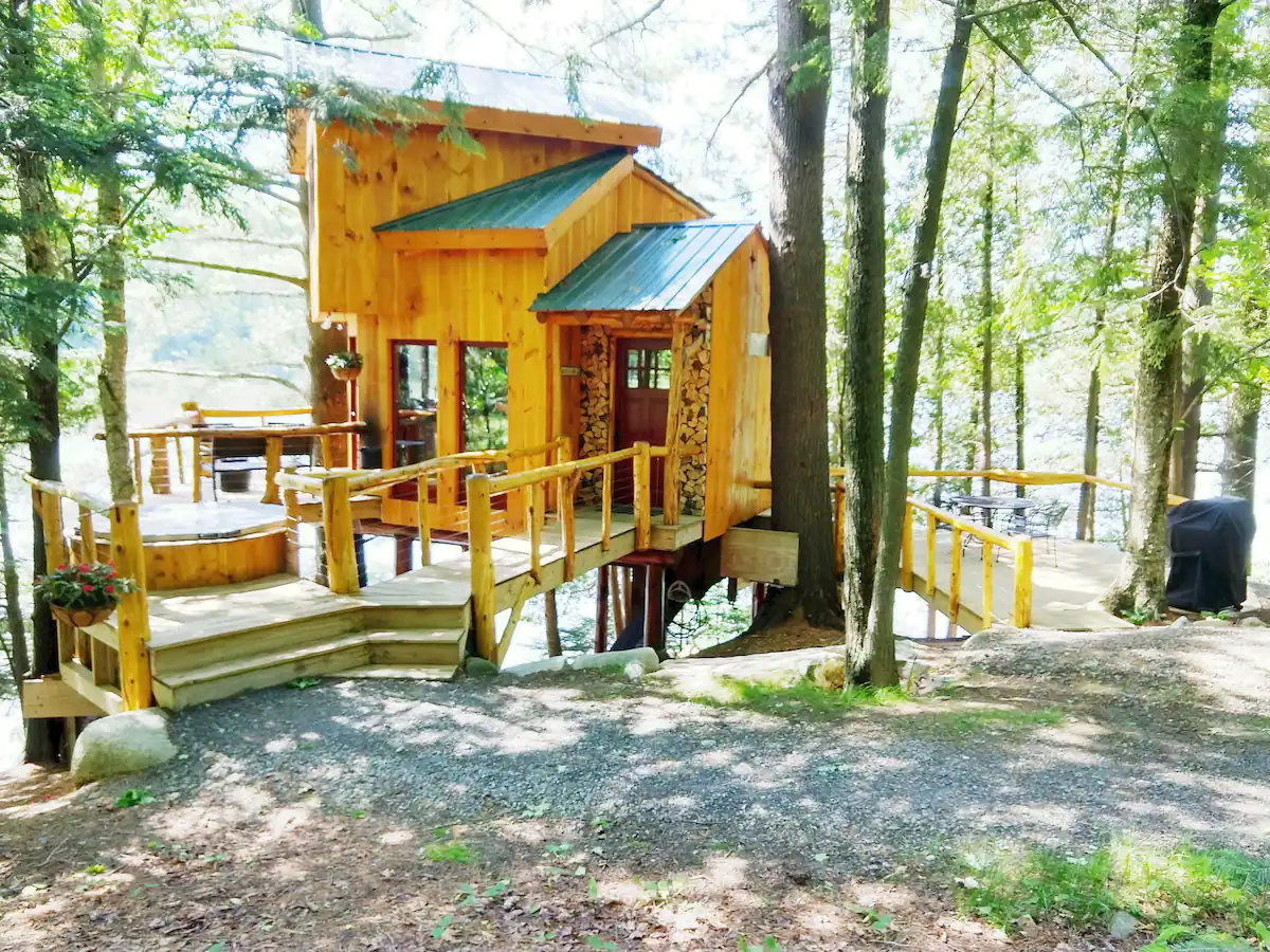 A wood cabin sits nestled in the woods with a hot tub on the deck and sunlight streaming through the trees