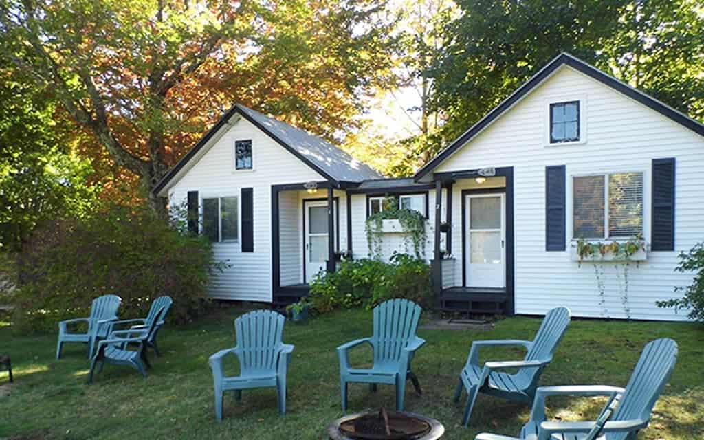 A firepit and aqua blue Adirondack chairs around it with a white cabins with blue trim behind them