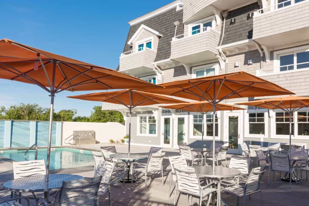 Tall white building with a patio next to pool with white furniture and red umbrellas