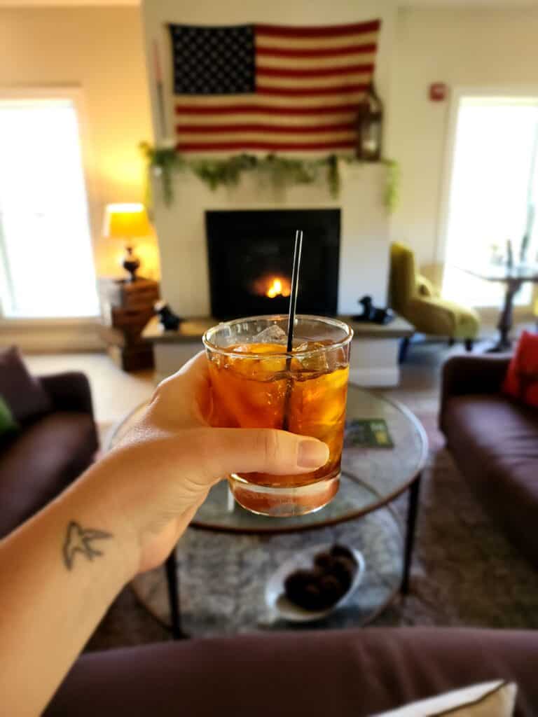 a hand holds a cocktail in the foreground, the background is a fireplace with an american flag above it. new england style