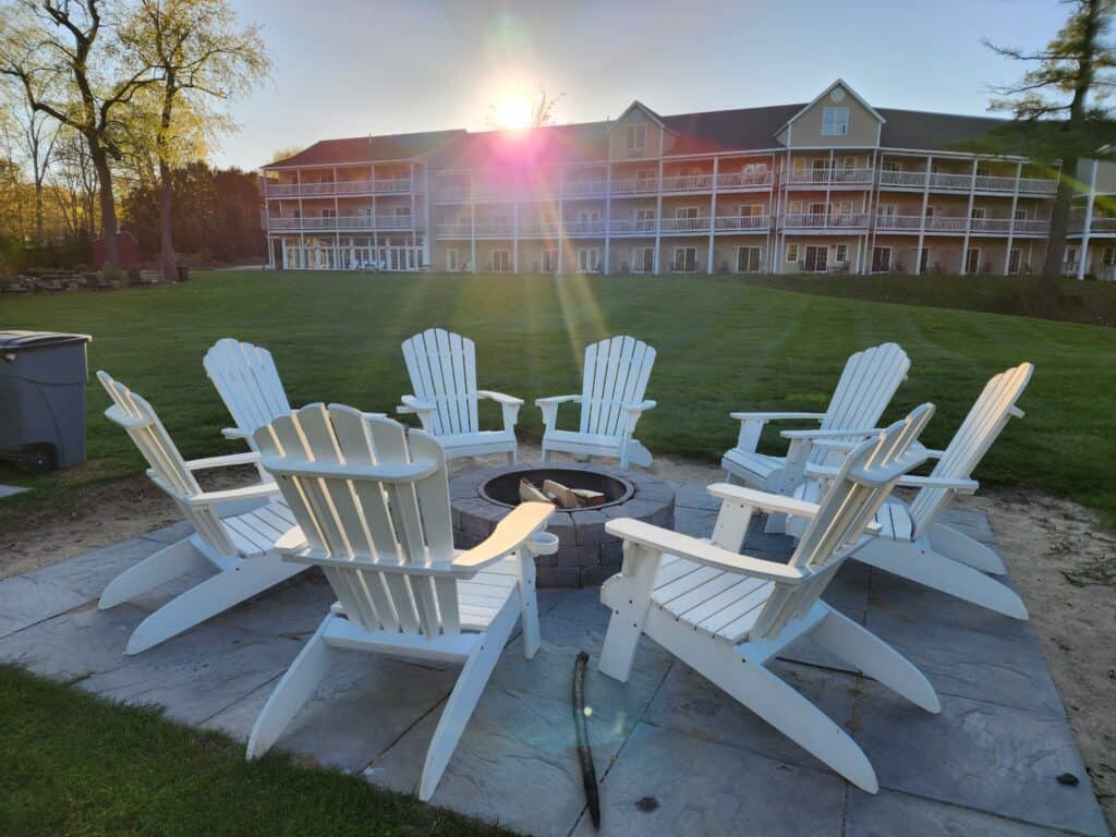 white adirondack chairs surround a firepit on the lawn behind a hotel. the sun is setting and flares over the scene. summer