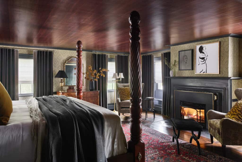 A dark wood four post bed sits surrounded by luxury antique furnishings as a fire roars in the fireplace