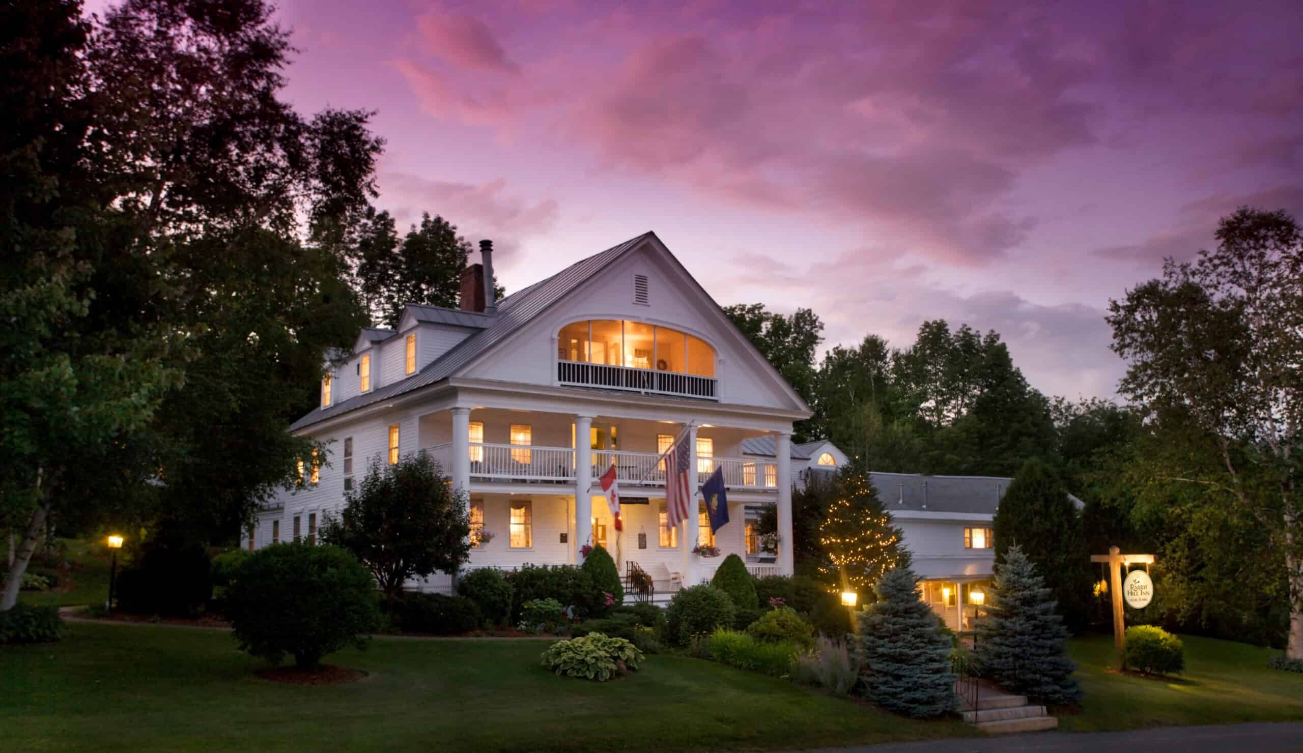 Large white house glowing with interior lights at dusk with a purple sky