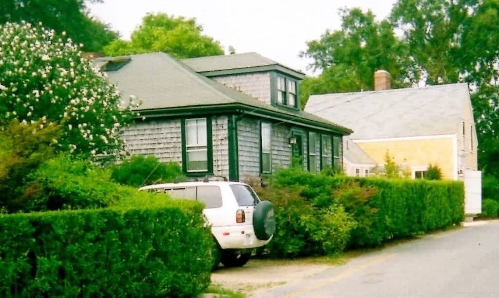 A grey Cape Cod house with green trim. Outside are many bushes and a white SUV.