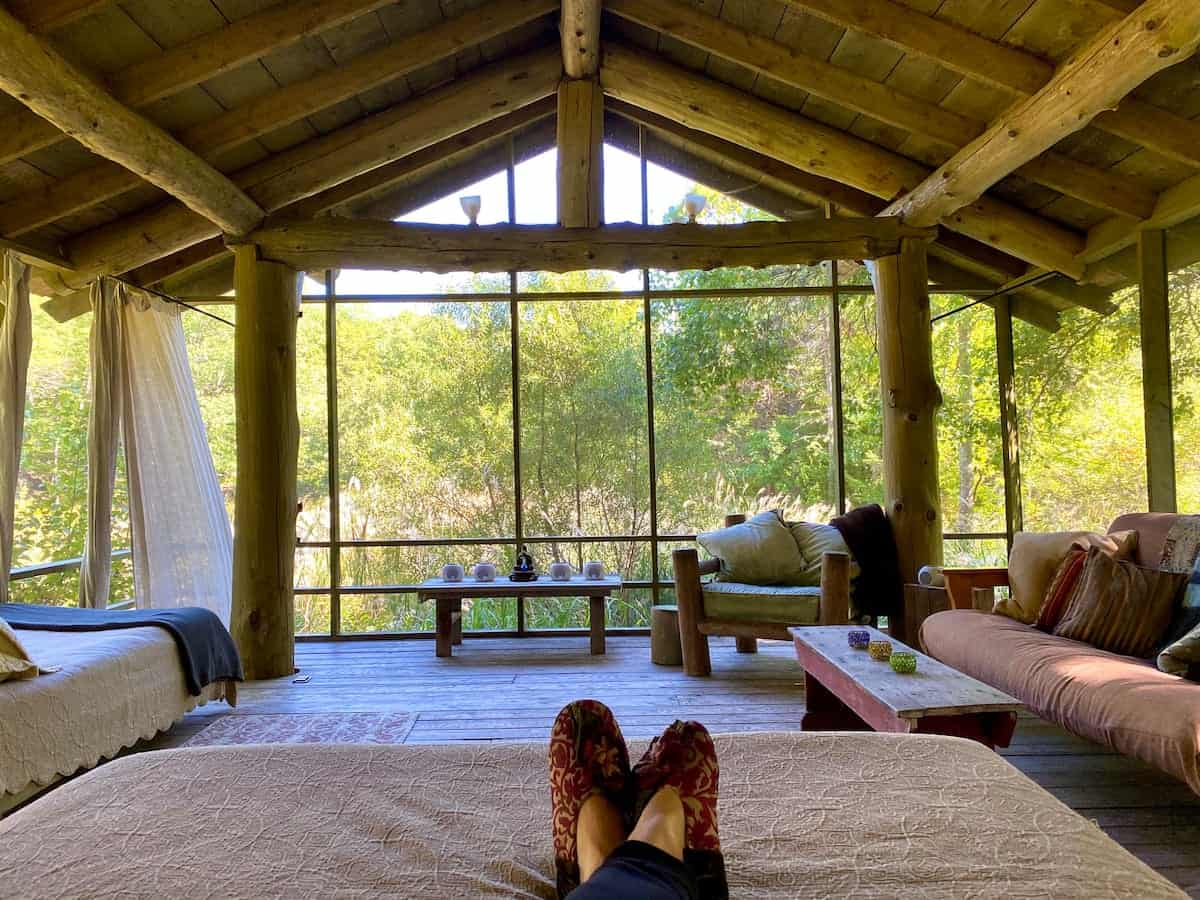 A woman\'s feet in slippers resting on a bed and facing a very large window that takes up the whole wall and looks out to shrubbery and trees at an off the grid Massachusetts cabin.