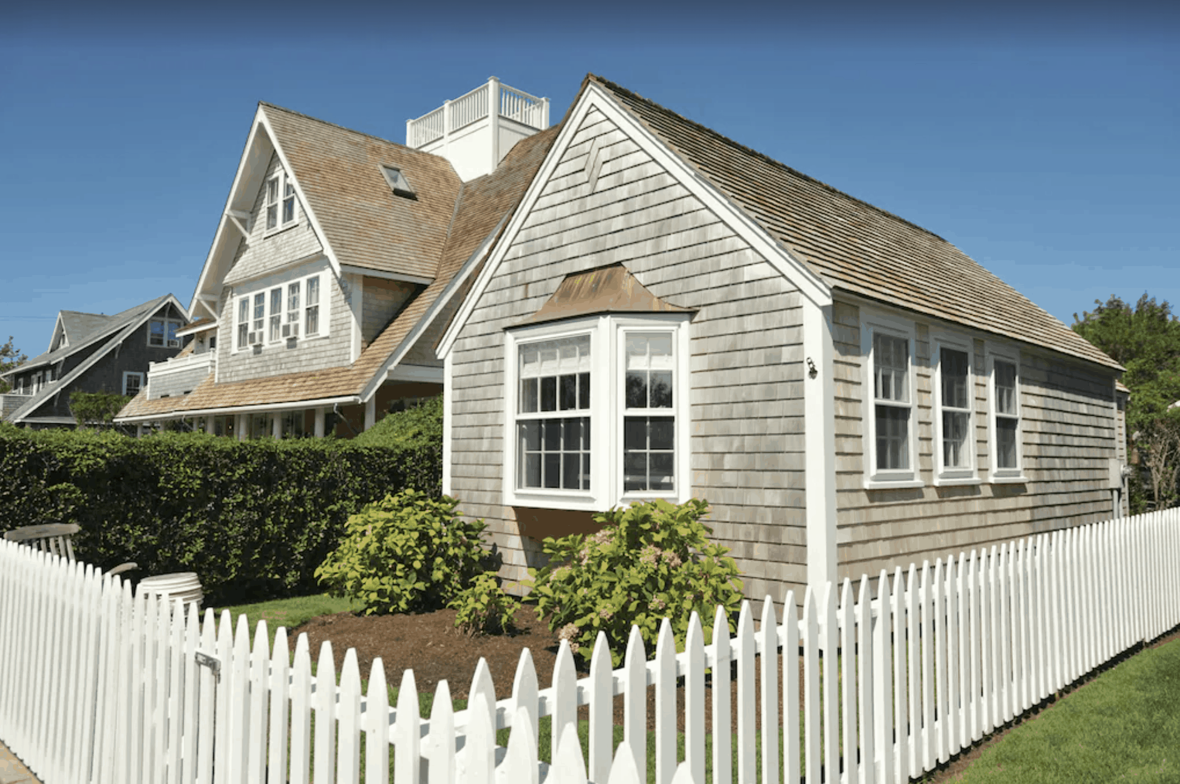 A grey cottage with a white picture window, surrounded by a white picket fence.