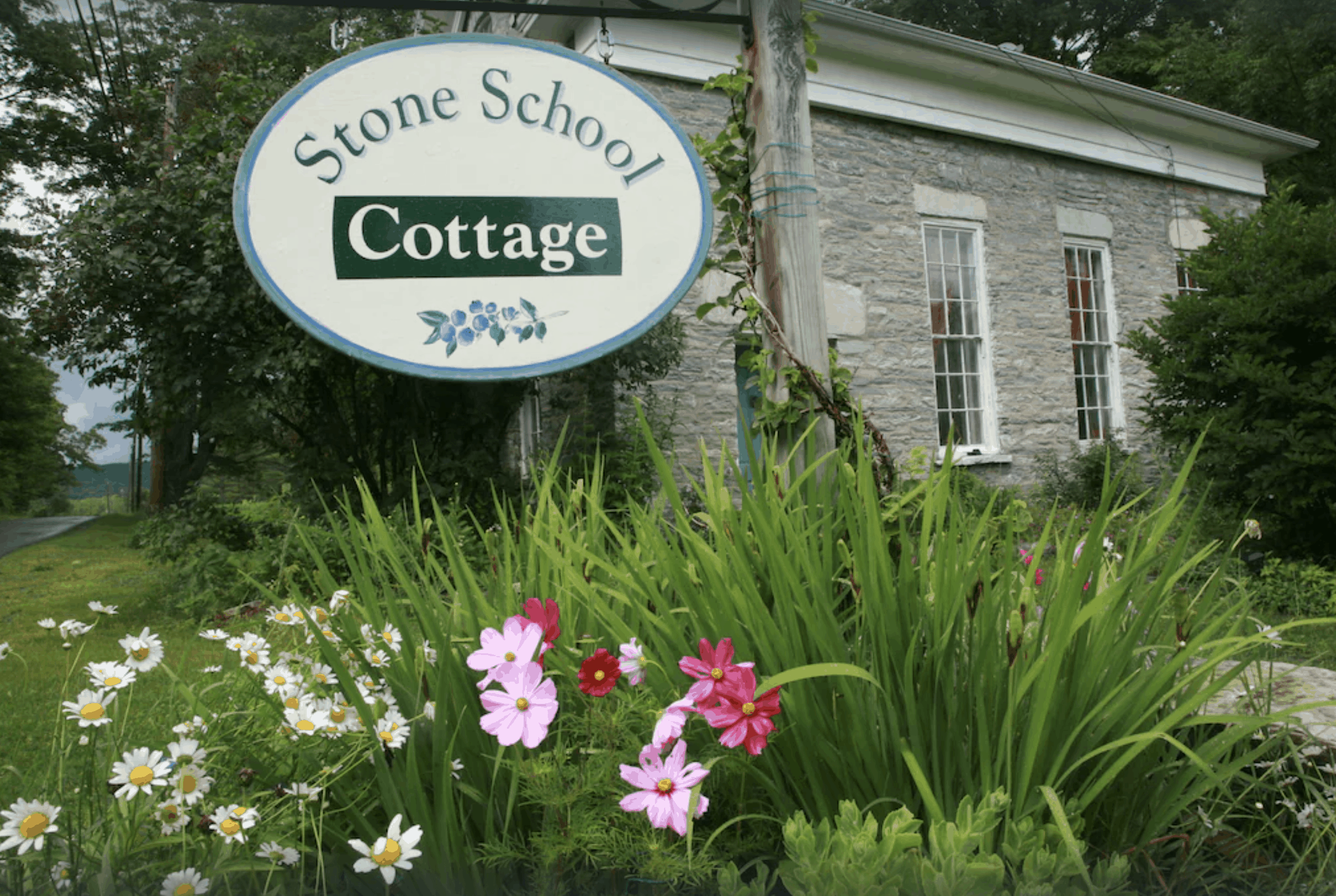 A grey cottage in Massachusetts with flowers out front, and a sign reading \"Stone School Cotttage\"
