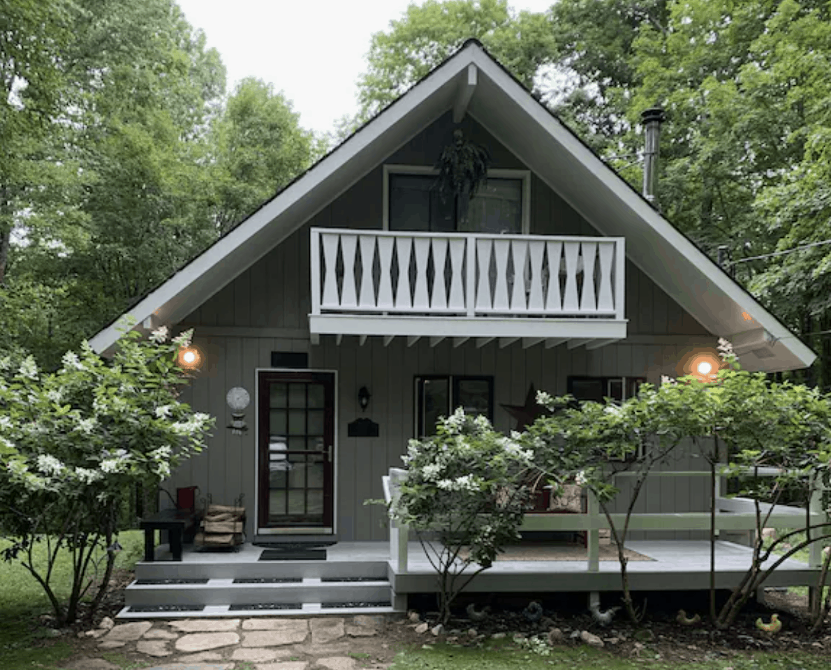 An A-Frame house with a white wooden balcony on the second floor, and a porch on the first floor. Surrounded by trees.