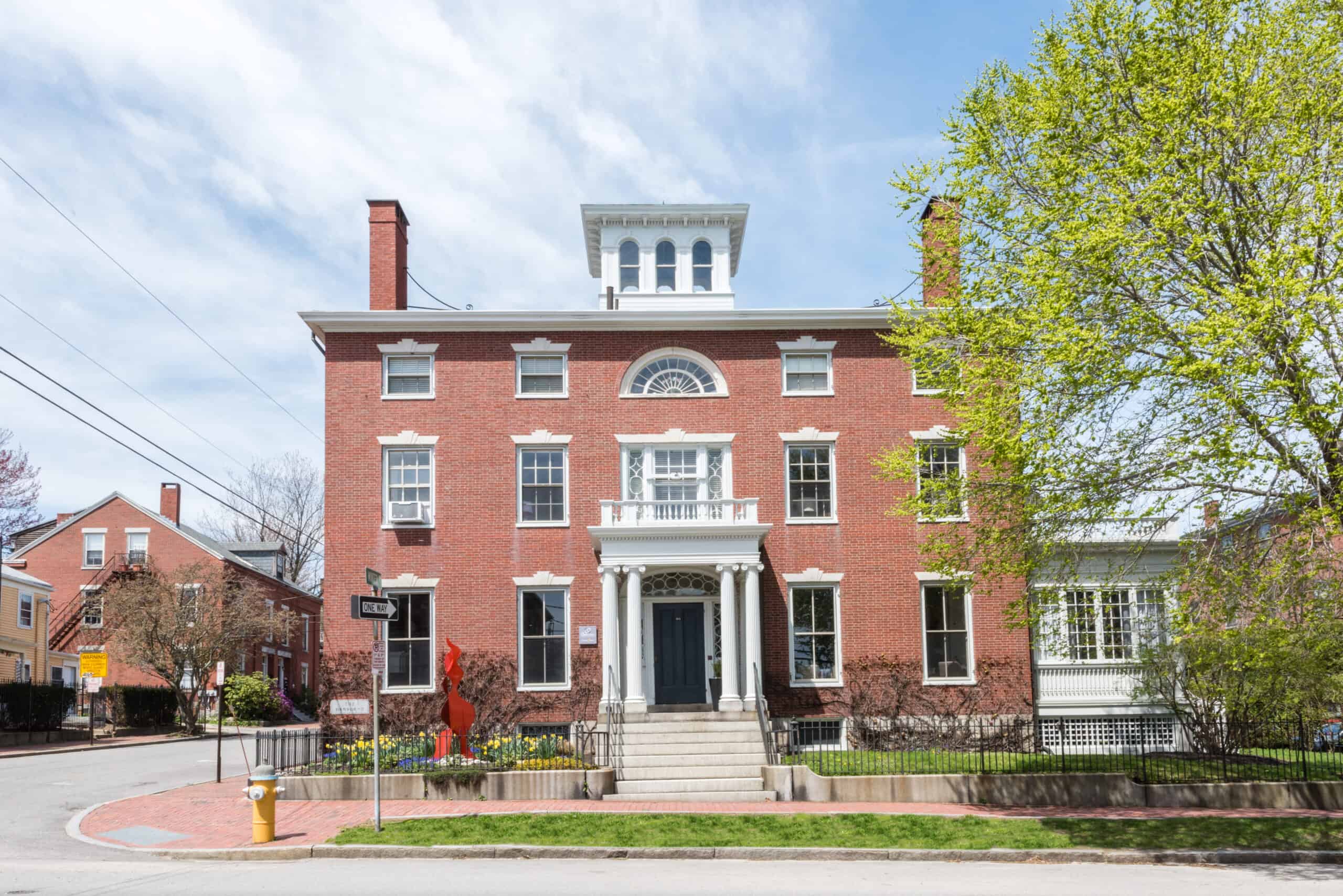 A stately red brick historic hotel in Portland Maine has three stories and is well landscaped with green grass and plants under a pale blue sky.