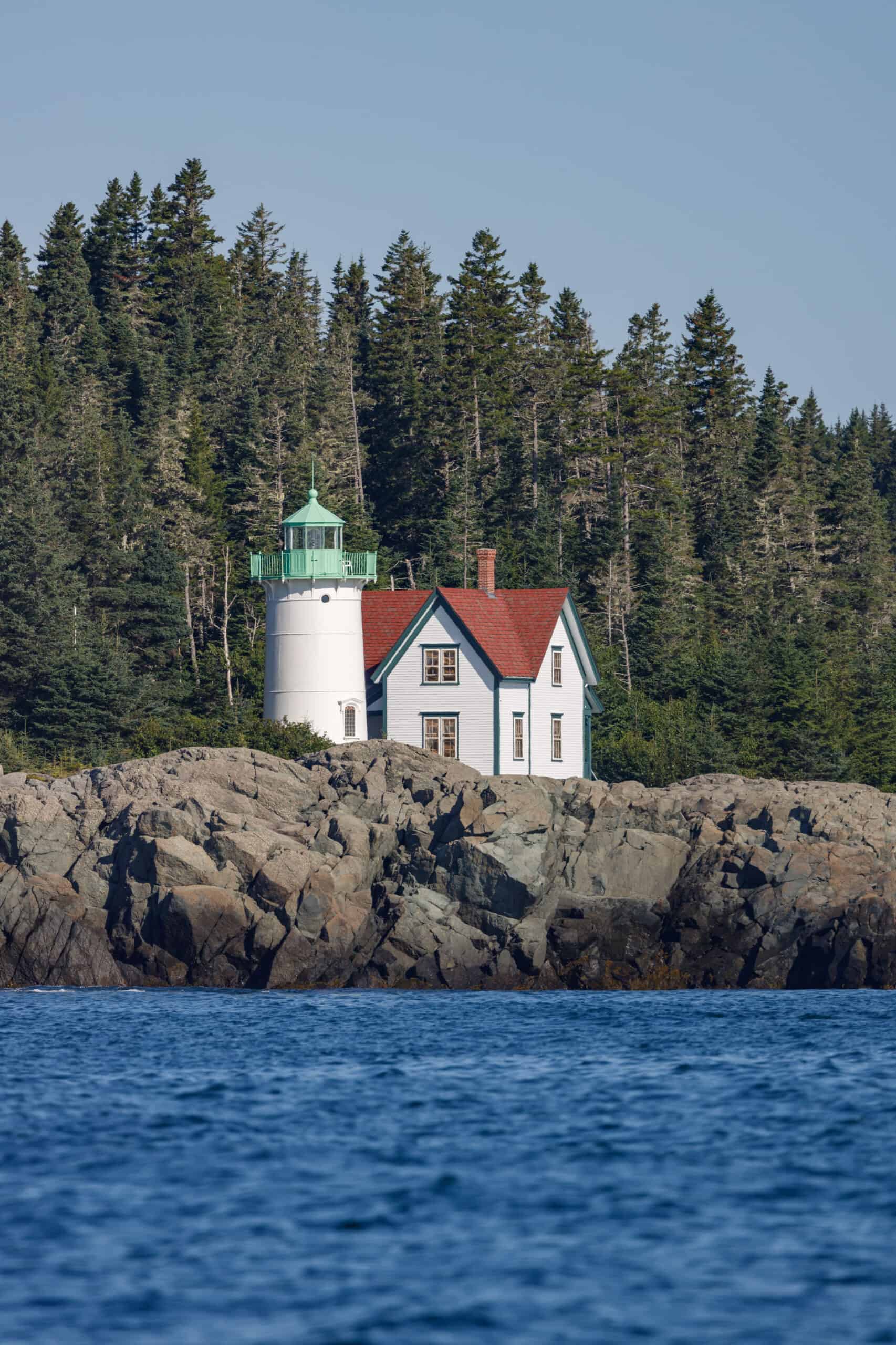 Lighthouse and lighthouse keepers house on rocky island in Maine with tall evergreen trees and blue skies behind it