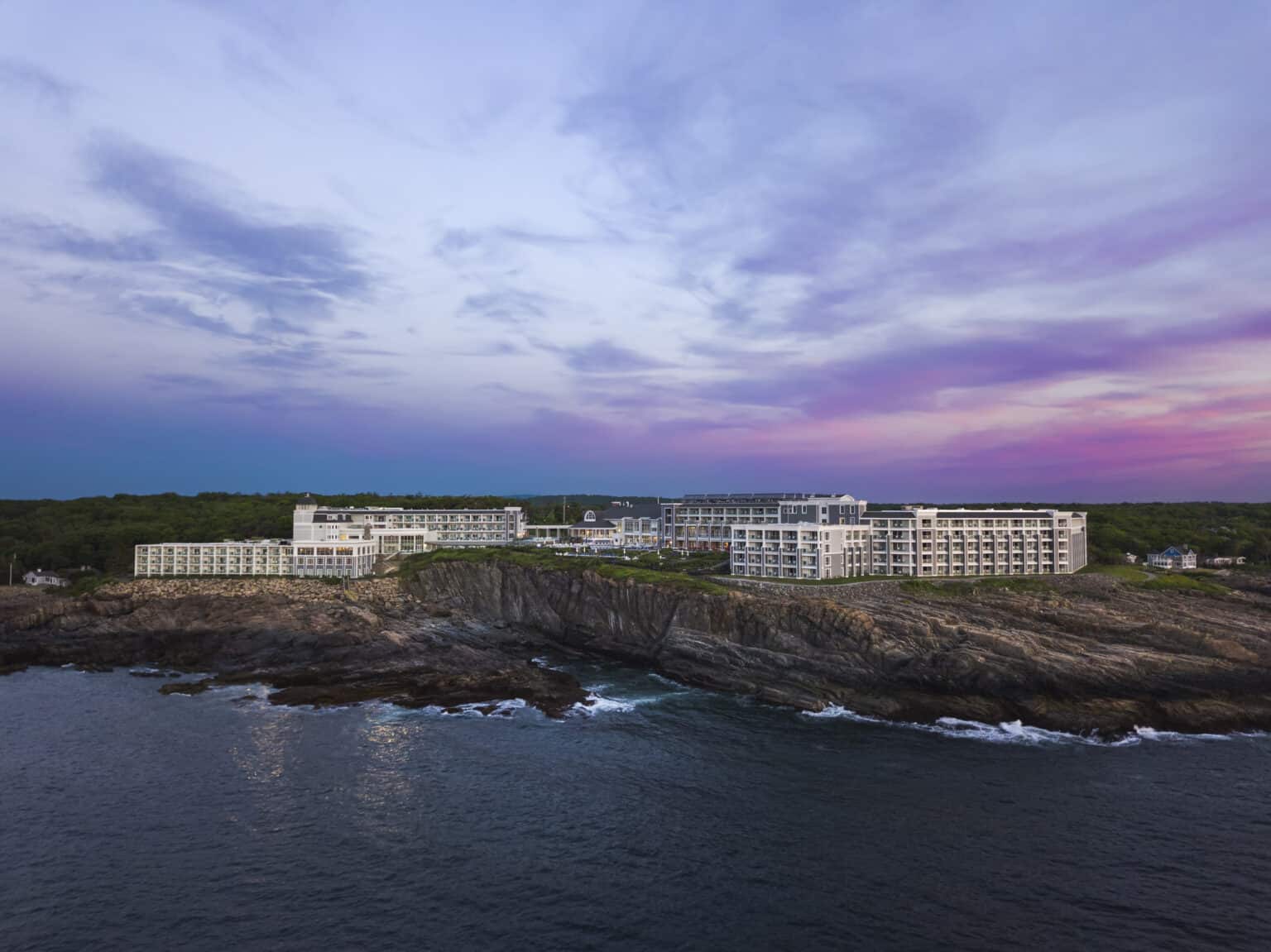 Sprawling white Cliff House resort perched dramatically on a cliff with the ocean below and a purple and pink sunset over head