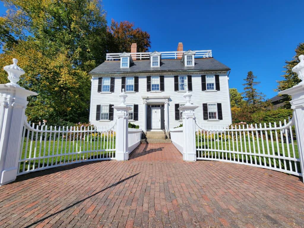 a white colonial style home set behind a broad white gate entrance. it is a sunny day with light fall foliage behind the house