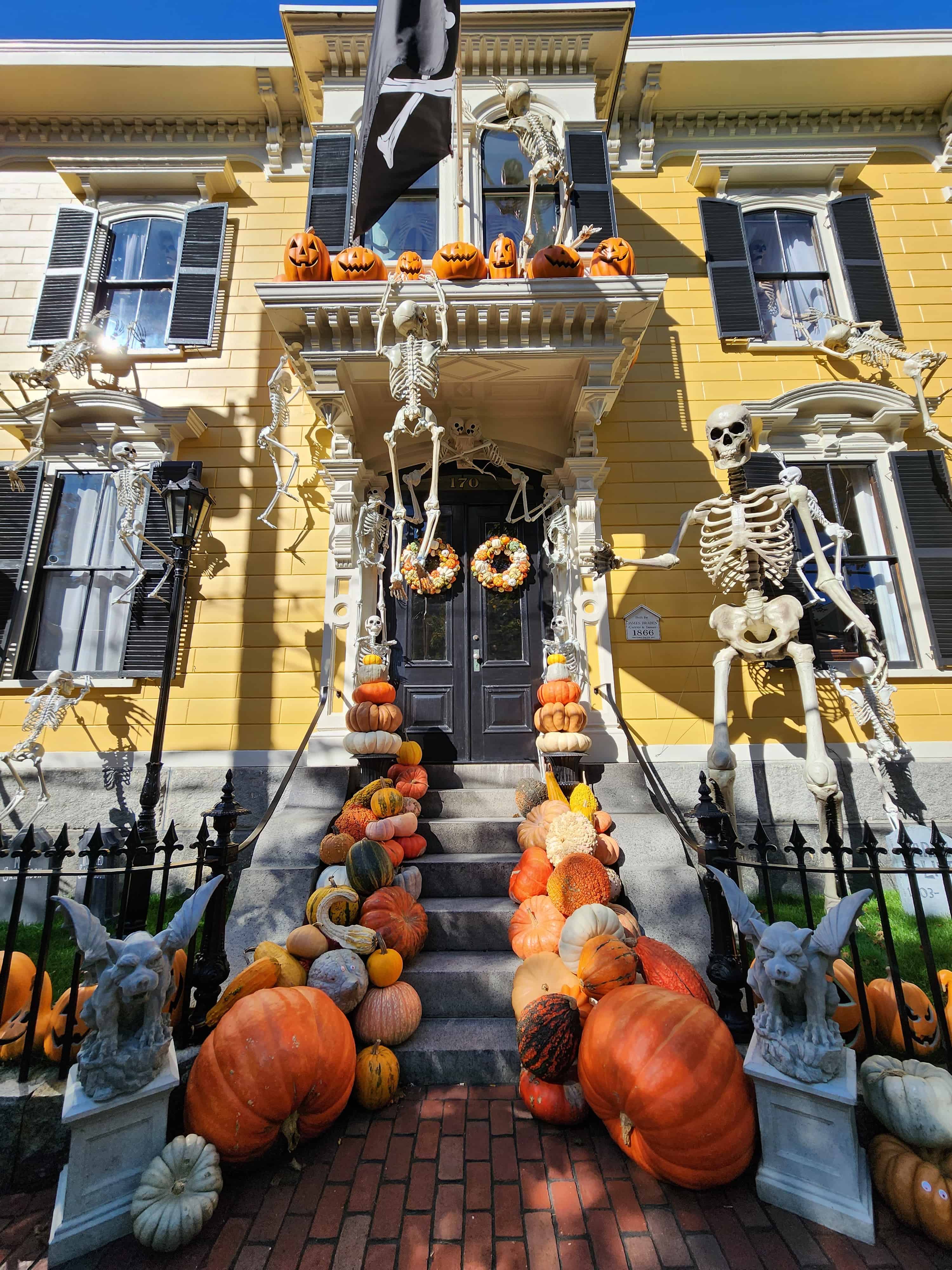 a yellow colonial home decorated for halloween with giant skeletons and lots of pumpkins