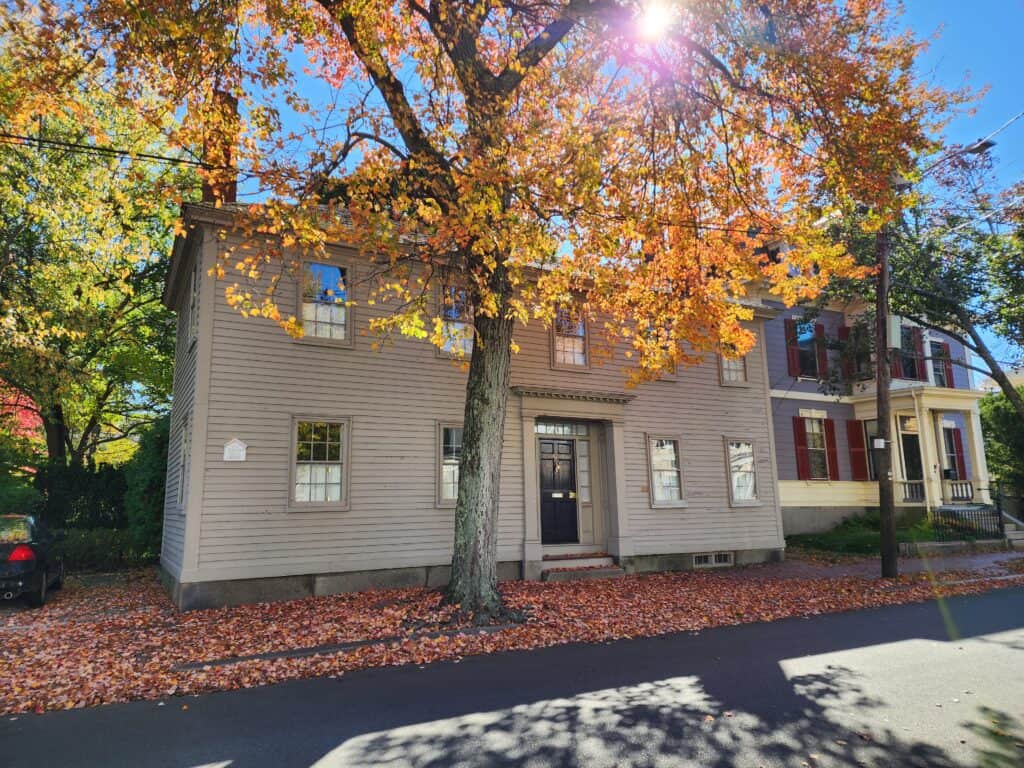 a grey older home behind a brightly colored fall tree with the sun shining through the branches