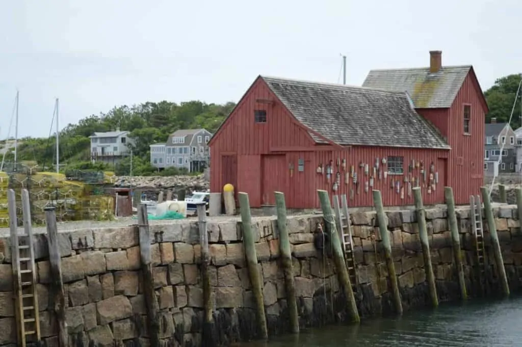 A red fishing shack is seen on a long stone pier with a coastal New England town beyond it