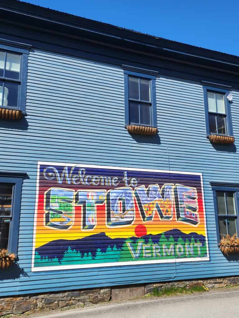 a colorful mural on the exterior wall of a blue house that looks like a vintage postcard and reads Welcome to Stowe Vermont