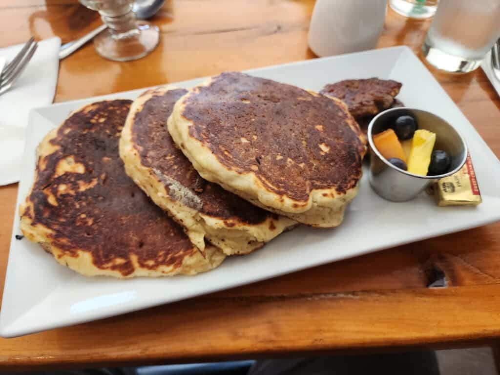 a side-lying stack of 3 fluffy pancakes is on a plate, with a small dish of fruit to the side.