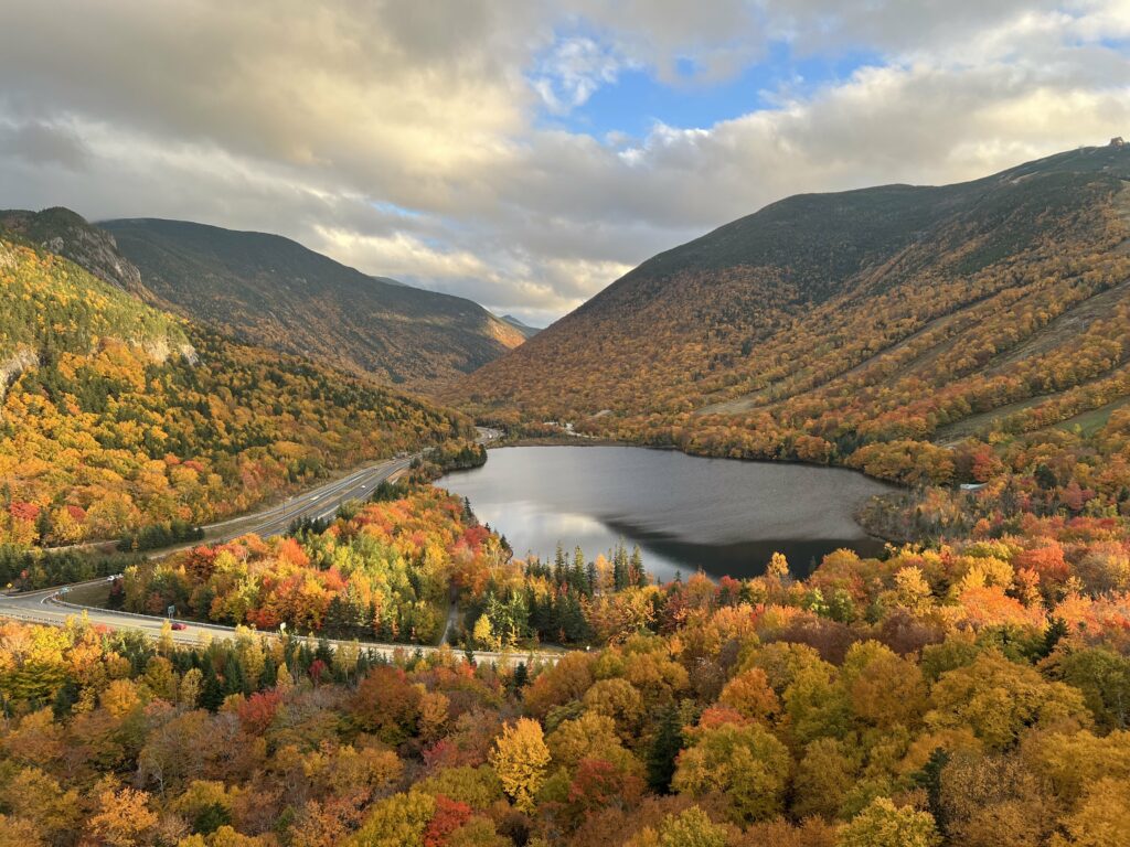 A panoramic view of one of the best spots in New England for fall, showcasing a serene lake surrounded by hills ablaze with the warm hues of autumn foliage, with a highway winding through the vibrant landscape