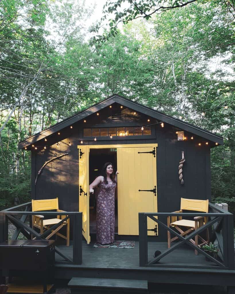 amy peeking out the door of a glamping cabin in maine - a brown cabin with yellow doors and fairy lights on the eaves