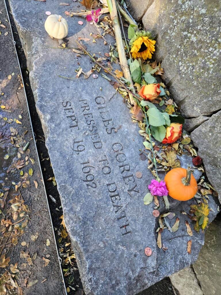 large stone slab with flowers, leaves, and a pumpkin. a carving reads: "Giles Corey Pressed to Death Sept. 19, 1692