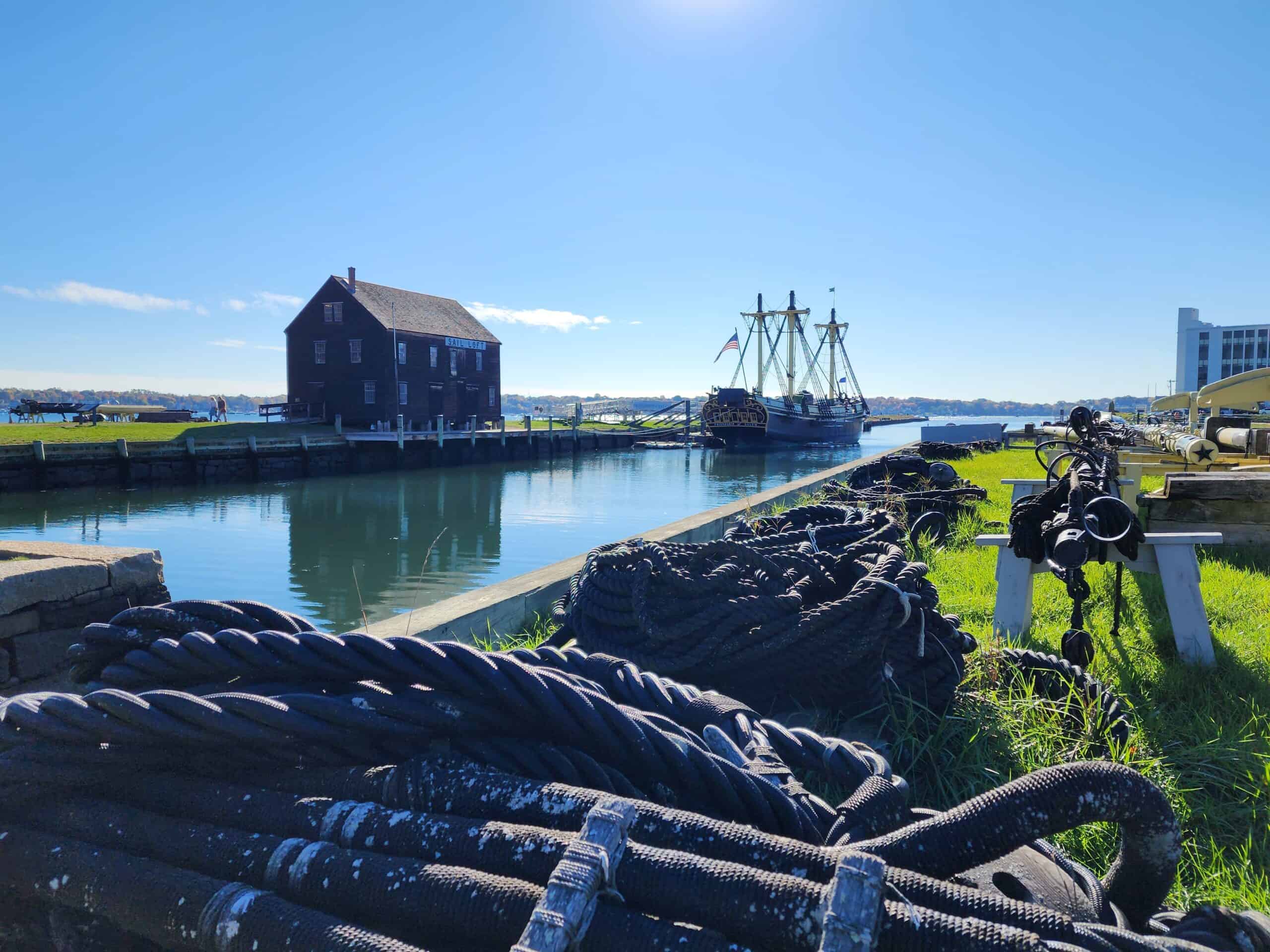 a stately colonial building abuts a calm harbor, where a tall masted sailing ship is anchored