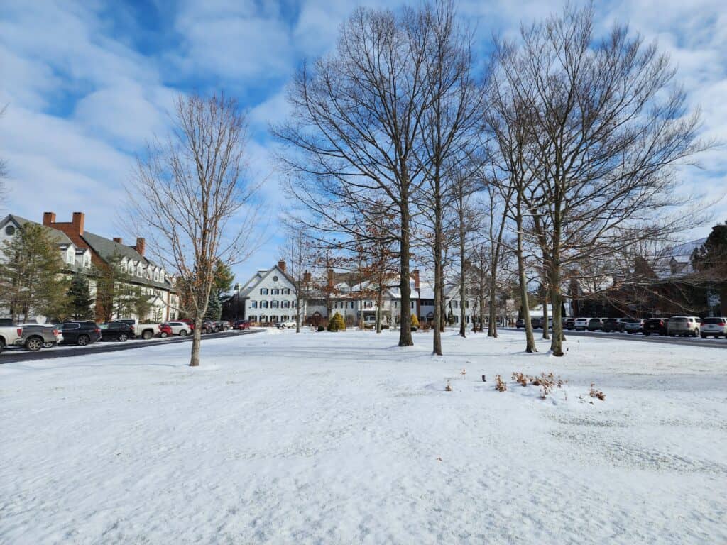 a snow covered field with a cozy inn in the distance behind some bare trees, winter in vermont