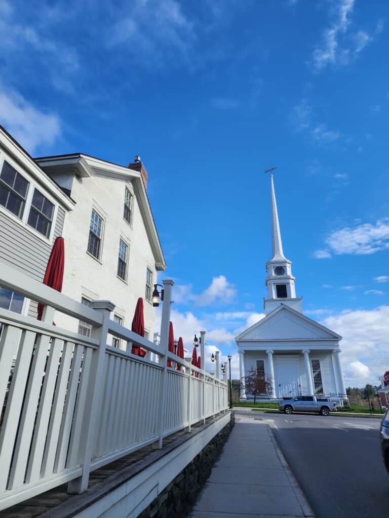 view from the end of a lane along a white picket fence, a white house to the left with a white steepled church in the distance
