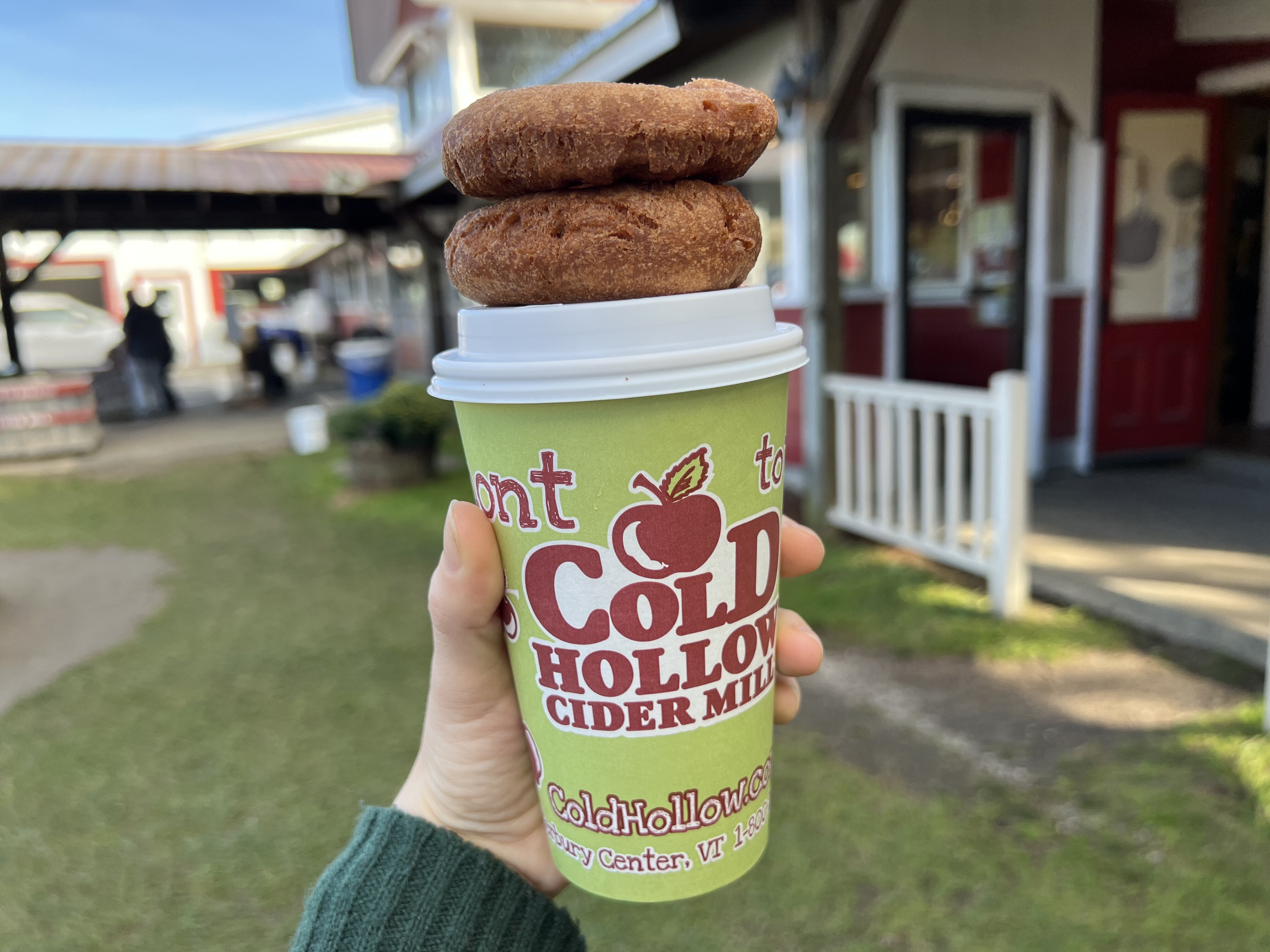 a hand holds a green coffee cup that reads cold hollow cider mill. atop the cup's lid are two stacked cider donuts. a blurry barn in the background