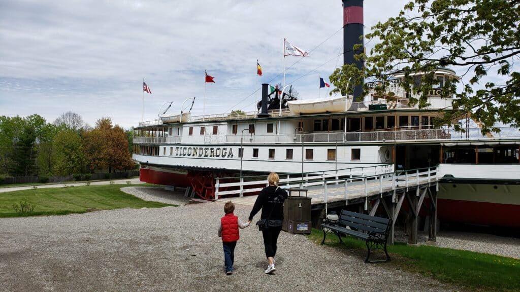 a woman and a young boy walk hand in hand towards a historic ship in an outdoor museum