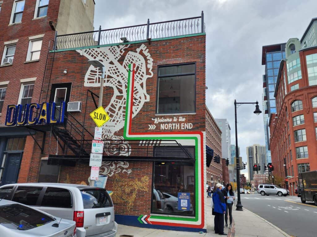 A mural of a bird on a brick building of one of the best North End restaurants Boston has to offer with traffic and people around