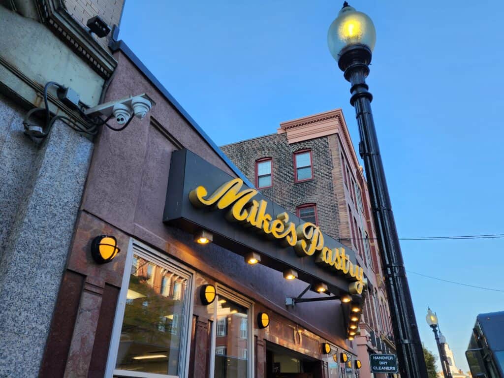 exterior of mike's pastry restaurant in boston north end. the sign is lit up with warm yellow lights and brick buildings are seen above and behind