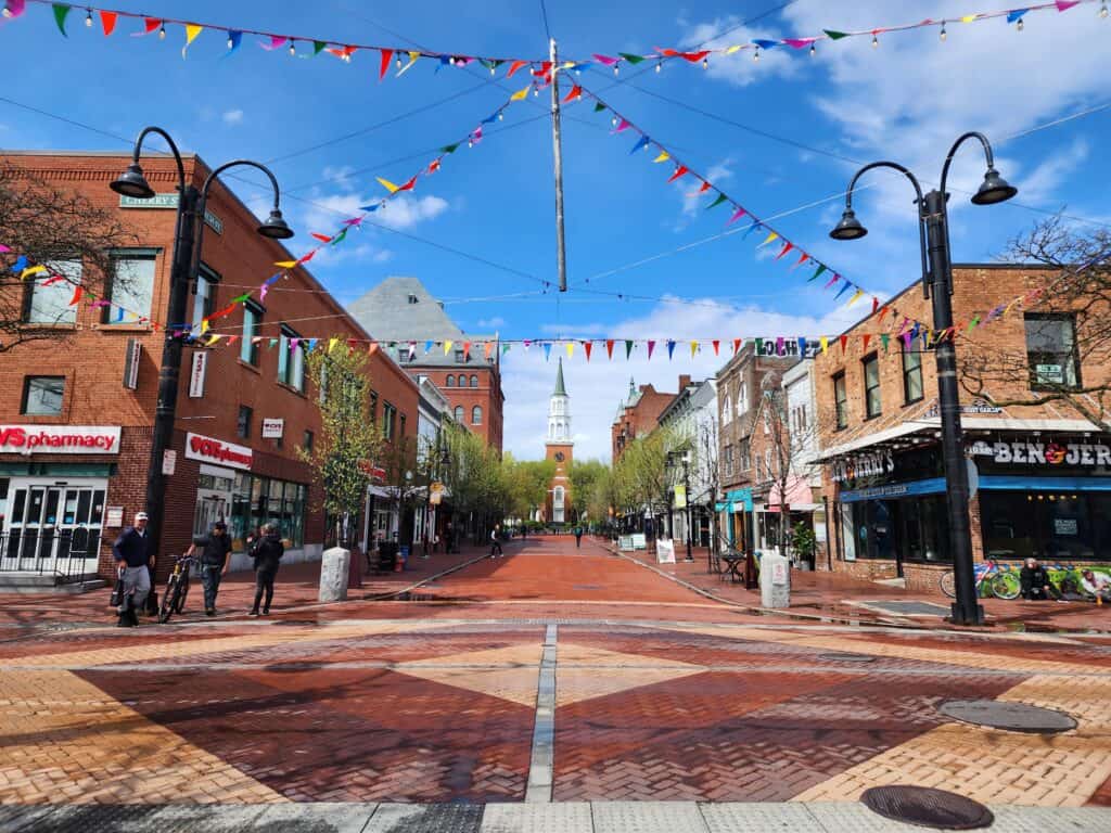an image of an empty shopping street on a sunny day. colorful banners cross over the blue sky and a long bricked walkway leads to a distant church - burlington vermont