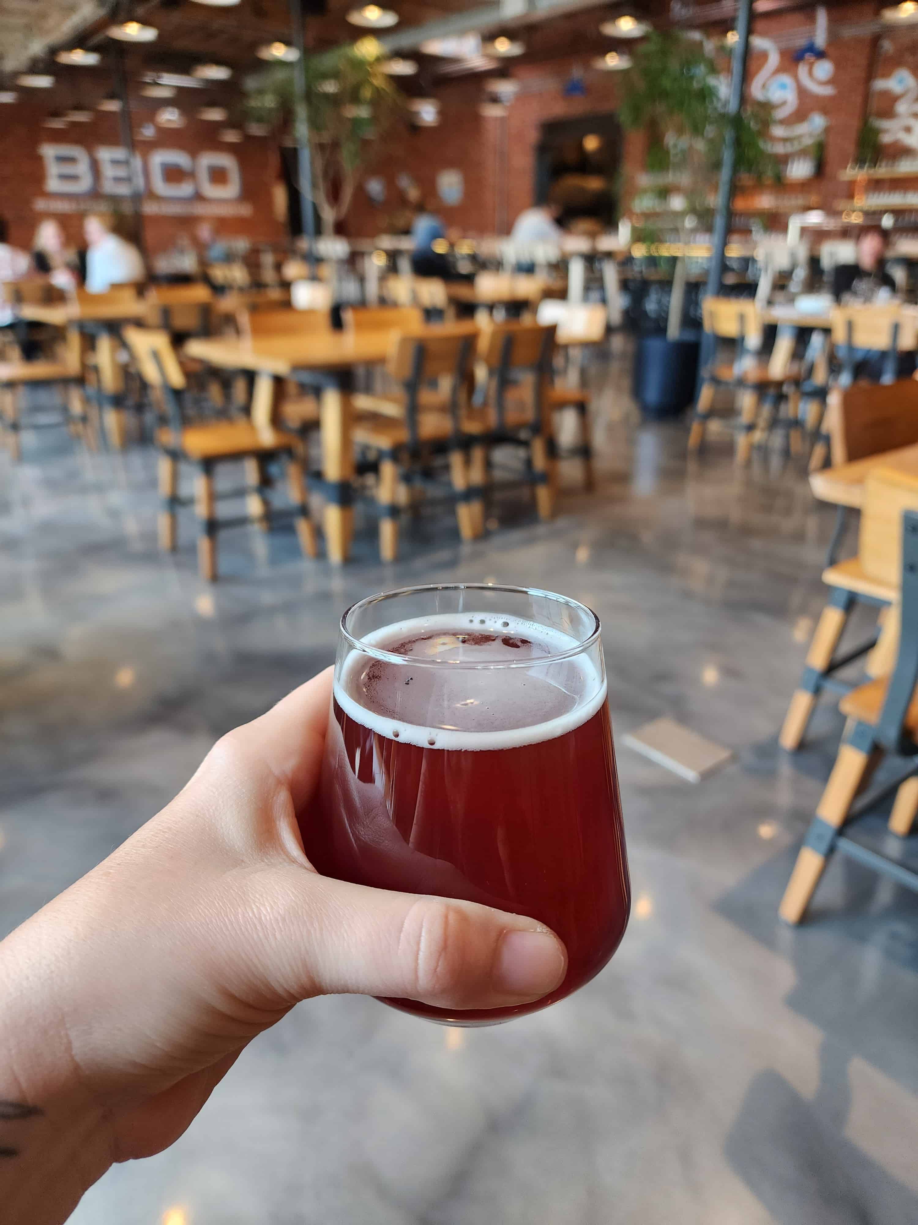 a hand holding a dark amber ale close to the camera, an out of focuse brewery restaurant is behind