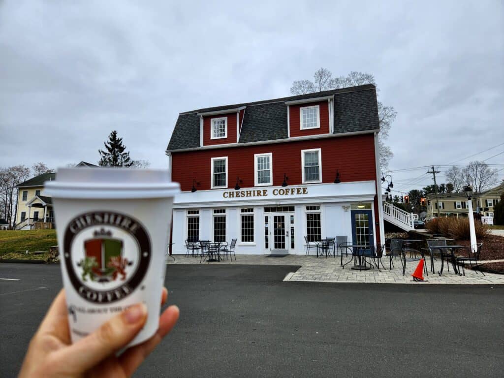 A hand holds a coffee cup up in front of one of the best coffee shops near New Haven Connecticut under grey skies