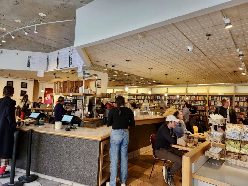A busy New Haven coffee shop is full of customers ordering coffee at a counter and sitting at tables with bookshelves along the walls