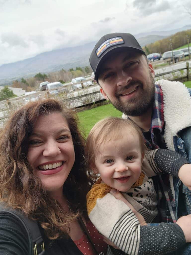 A man, woman, and toddler smile in front of scenic mountains in Stowe, Vermont