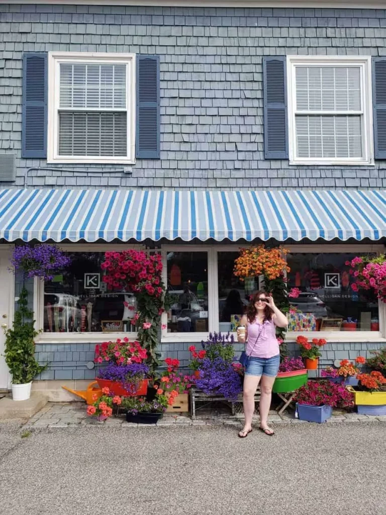 A girl stands in front of a store with flowers surrounding her with a blue and white awning.