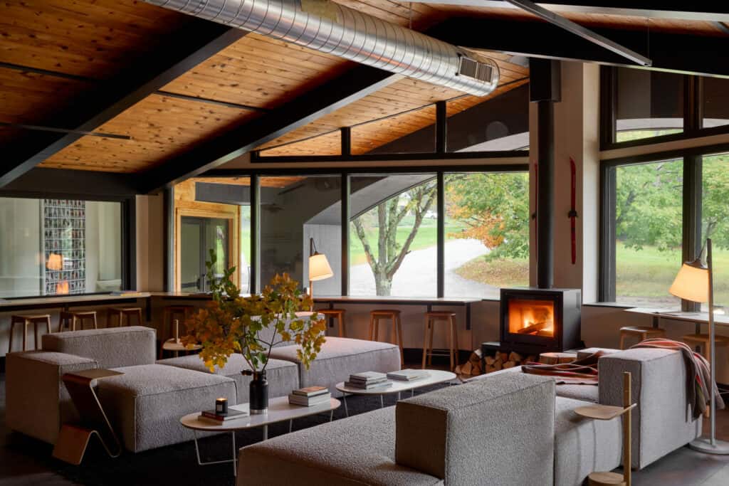 interior image of a hotel, a chic cabin-inspired gathering space featuring large windows, a vaulted wooden roof, a simple woodstove, and understated grey couches