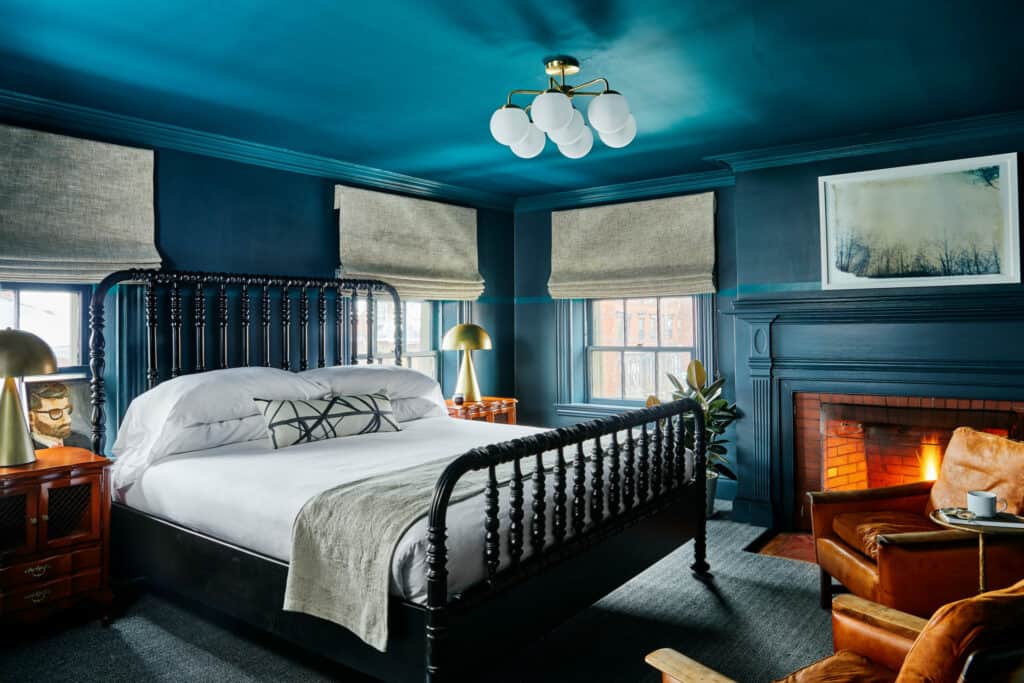 A room in one of the most popular boutique hotels Portland Maine has to offer features deep blue walls, ceiling, and mantle, with a bed in the center and a fire roaring in the fireplace