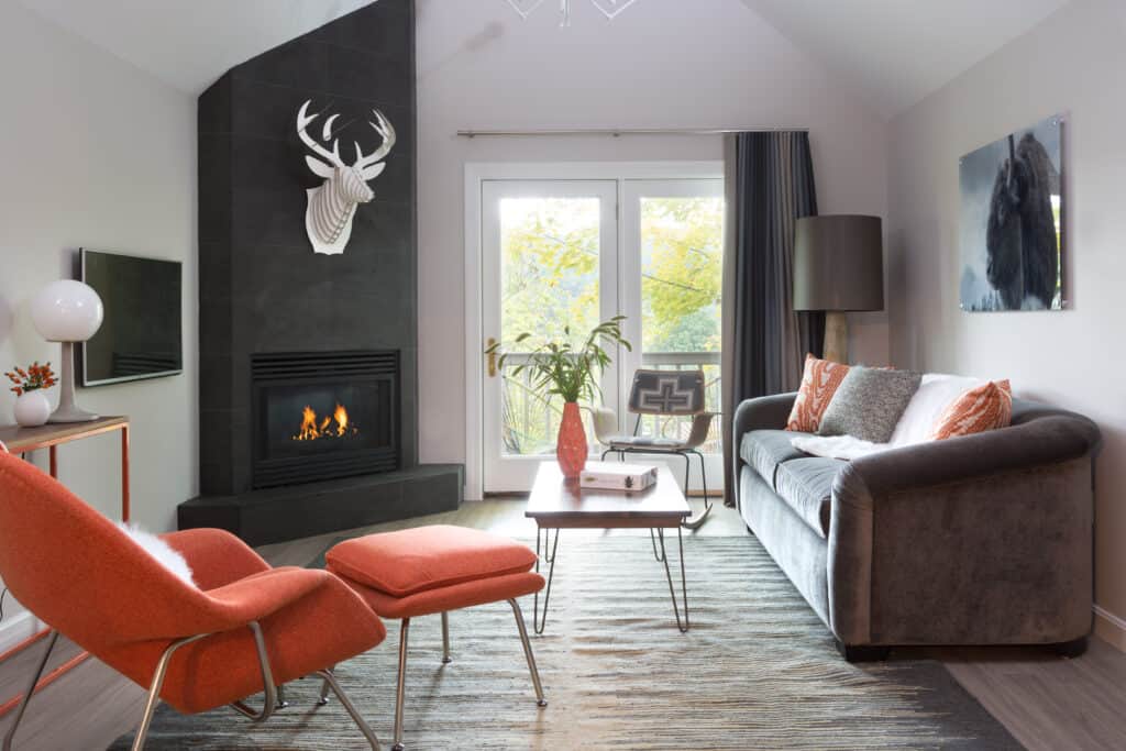 cozy and modern hotel room interior - a living room setup with grey and coral furniture, a lit fireplace is topped with an all white deer bust